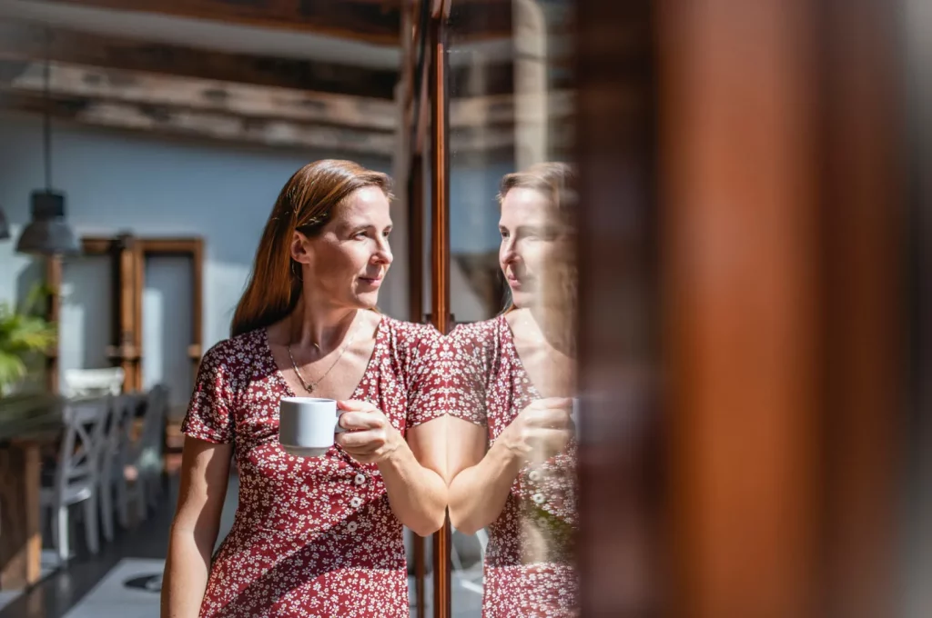 Mujer reflexiva con taza de café mirándose al espejo, representando el inicio de un viaje de autoconocimiento y pausa emocional